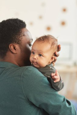 Afro-Amerikalı bir adamın dikey arka plan portresi. Elinde şirin melez bir bebek var. Evinde rahat bir şekilde poz verirken kameraya bakıyor.