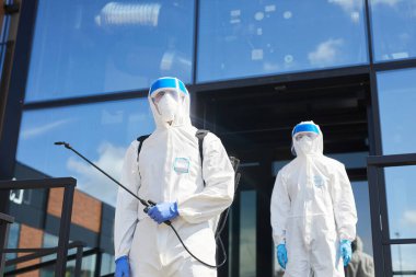 Low angle view at two workers wearing protective suits posing with disinfection gear outdoors while standing against glass building, copy space clipart