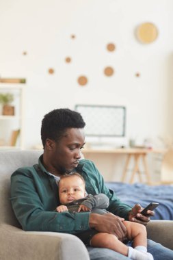 Genç Afro-Amerikan babanın dikey portresi. Kucağında şirin melez bir bebek, rahat koltukta oturuyor ve akıllı telefon kullanıyor, kopyalama alanı kullanıyor.