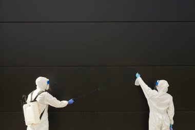 Wide angle view at two workers wearing protective gear and spraying chemicals over black building facade during disinfection or cleaning, copy space clipart