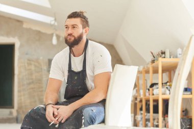 Yetişkin sakallı bir sanatçının ilham verici portresi. Güneş ışığıyla aydınlatılmış bir sanat stüdyosunda otururken dalgın dalgın bakıyor ve sigara içiyor.