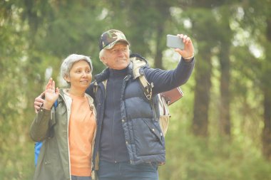 Aktif kıdemli çiftin belden yukarı portresi akıllı telefondan fotoğraf çekerken ormanda yürüyüş yapmak, uzay kopyalamak...