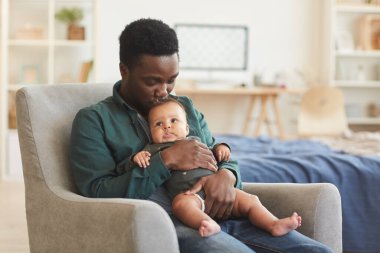 Evdeki rahat koltukta otururken tatlı melez bir bebeği tutan Afrikalı Amerikalı bir adamın portresi.