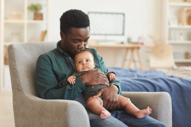 Portrait of caring African-American man holding cute mixed-race baby while sitting in comfortable armchair at home, copy space clipart
