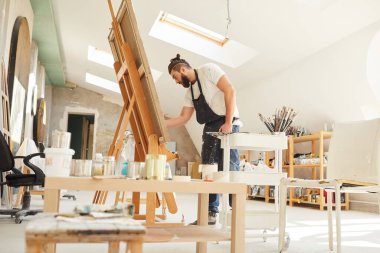 Günışığı ile aydınlatılan geniş bir sanat stüdyosunda dikilirken sehpada resim çizen yetişkin çağdaş sanatçıya geniş açı görünümü