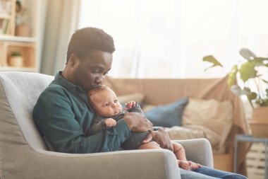 Evdeki rahat koltukta otururken sevimli melez bir bebeği tutan Afrikalı Amerikalı adamın yan görünüm portresi.