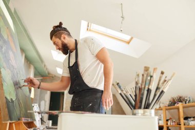 Çağdaş sakallı ressamın geniş tavan arası sanat stüdyosunda dikilirken sehpaya resim çizmesinin yan görünüş portresi.