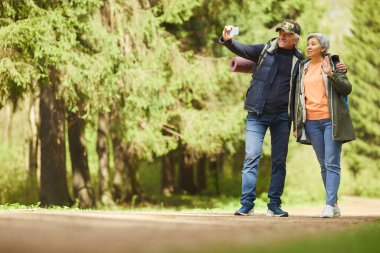 Ormanda yürüyüş yaparken akıllı telefondan selfie çeken aktif yaşlı bir çiftin tam boy portresi.