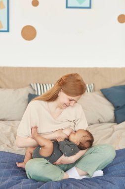 Gülümseyen yetişkin annenin dikey portresi melez bir bebeği emzirirken evde oturup, boşluğu kopyalıyor.