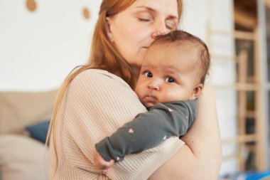 Sevgi dolu yetişkin annenin, evde anneliğin tadını çıkarırken tatlı melez bebeği kucaklamasının sıcak tonlu yakın plan portresi. Kopya.