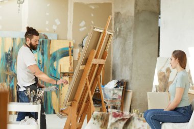 Stüdyoda genç bir kadının portresini yapan sakallı bir erkek ressamın yan görüntüsü.