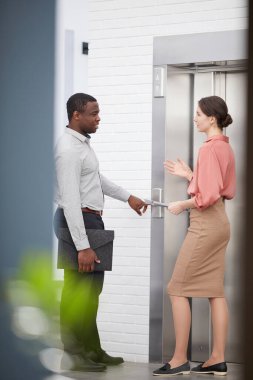 İki çağdaş ofis çalışanının tam boy portresi koridorda asansörü beklerken neşeyle sohbet ediyorlar.