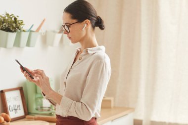 Zarif bir iş kadınının sıcak tonlu yan görünüm portresi akıllı telefon kullanırken rahat bir evde duruyor, boşluğu kopyalıyor.