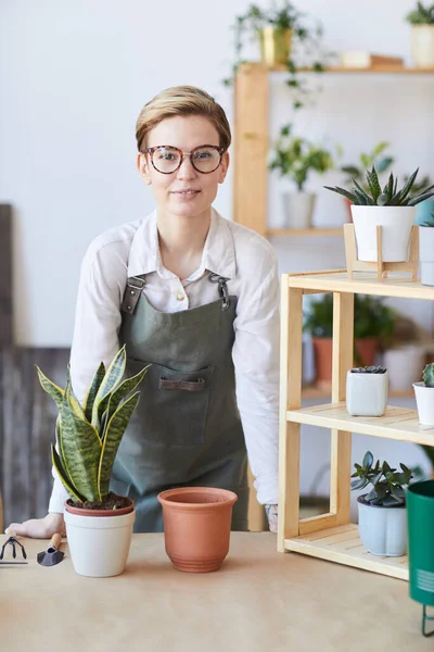 鉢植えや土壌で木製のテーブルに立ってエプロンを身に着けている現代の若い女性の垂直肖像画は 家庭菜園の概念の準備ができて — ストック写真