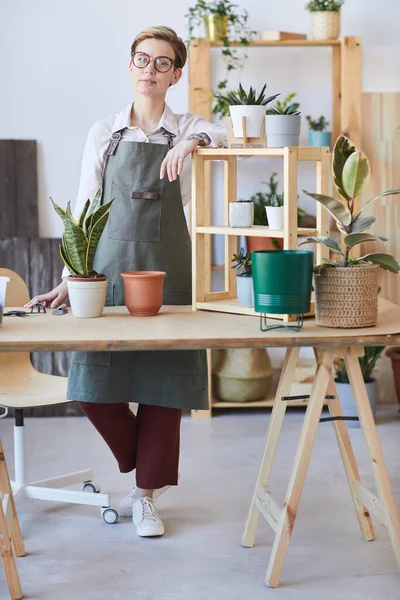 鉢植えや土壌で木製のテーブルに立ってエプロンを身に着けている現代の若い女性の垂直フル長さの肖像画は 鉢植え 家庭菜園のコンセプトの準備ができて — ストック写真