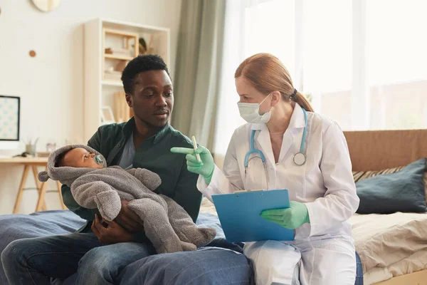 Portrait of young African-American dad holding cute baby and listening to female doctor during medical checkup at home, copy space
