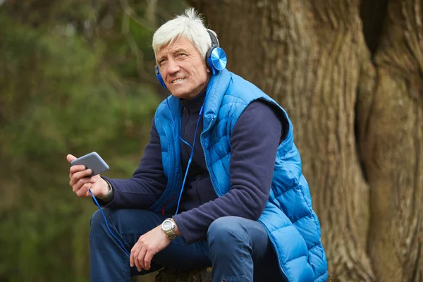 Portrait of active senior man listening to music via smartphone while sitting by tree and resting during hike in autumn forest, copy space