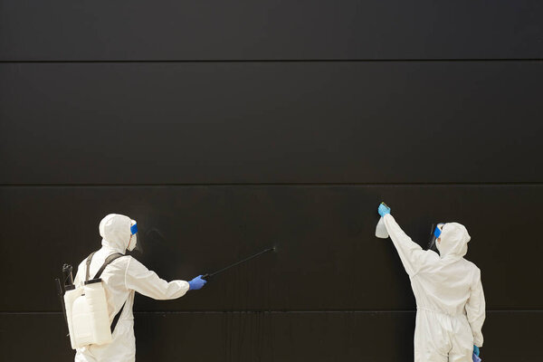 Широкий угол обзора на двух рабочих, носящих защитное снаряжение и распыляющих химикаты над черным фасадом здания во время дезинфекции или очистки, копирования пространства