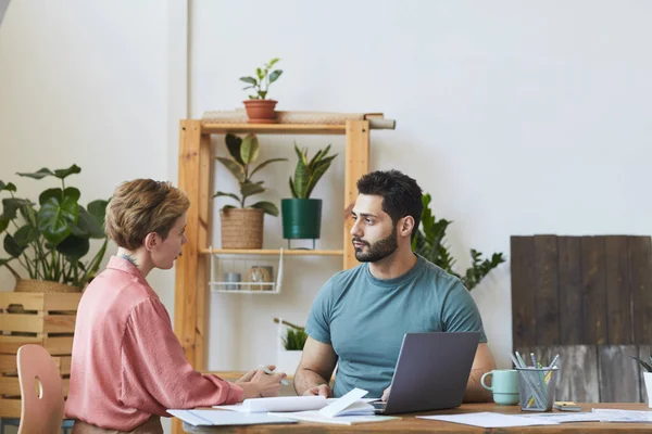 Portrait of handsome bearded man talking to female colleague while working at desk in modern office interior, copy space