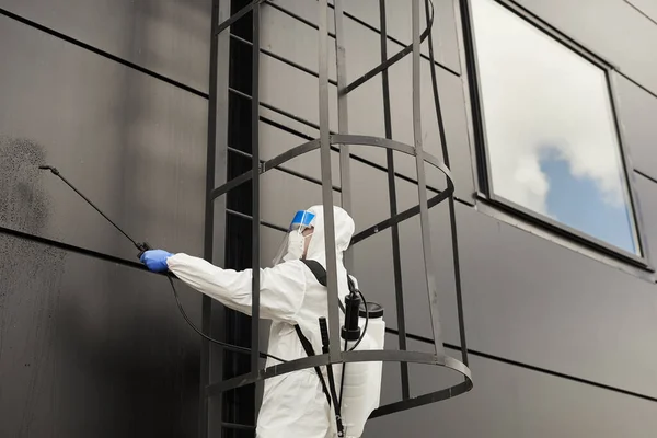 在黑色建筑物外墙进行消毒或清洁 复制空间时 一名工人穿著防护服在外墙喷洒化学品的广角视野 — 图库照片