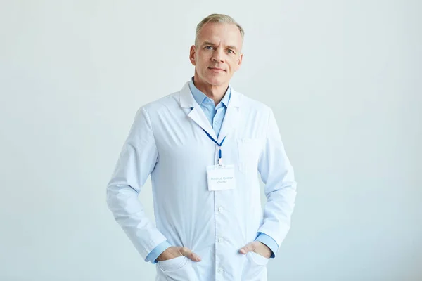 Waist up portrait of confident mature doctor wearing lab coat and looking at camera while standing against white wall in clinic, copy space