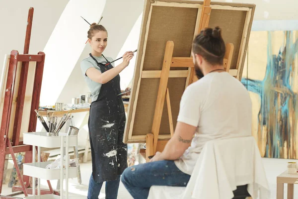 Portrait of young female artist painting portrait of man in studio, copy space