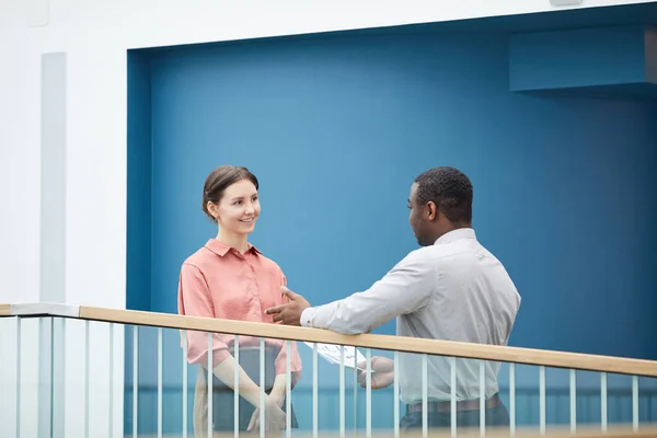 青い壁に対してバルコニーに立っている間 アフリカ系アメリカ人の同僚と話している若いビジネスマンの笑顔の肖像画をウエストアップ スペースをコピー — ストック写真