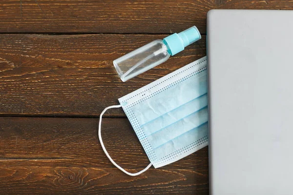 Close up of laptop, face mask and hand sanitizer over dark wooden background, quarantine home office workplace, copy space