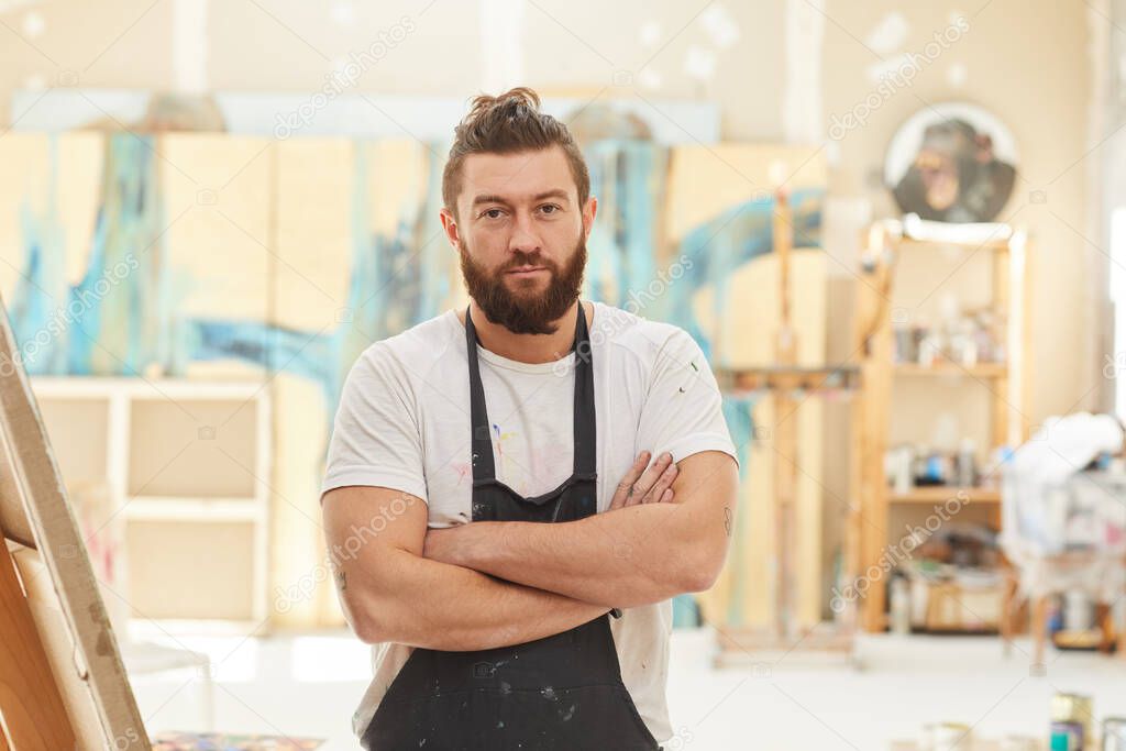 Waist up portrait of mature bearded artist standing with arms crossed and looking at camera while posing in spacious art studio lit by sunlight, copy space
