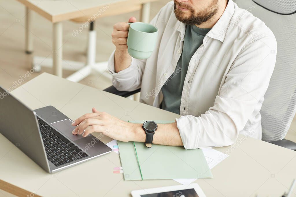 High angle view at bearded adult man drinking coffee while using laptop workplace in minimal interior, copy space