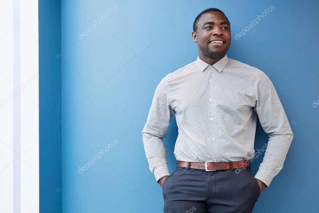 Waist up portrait of successful African-American businessman looking away and smiling while leaning against blue wall, copy space