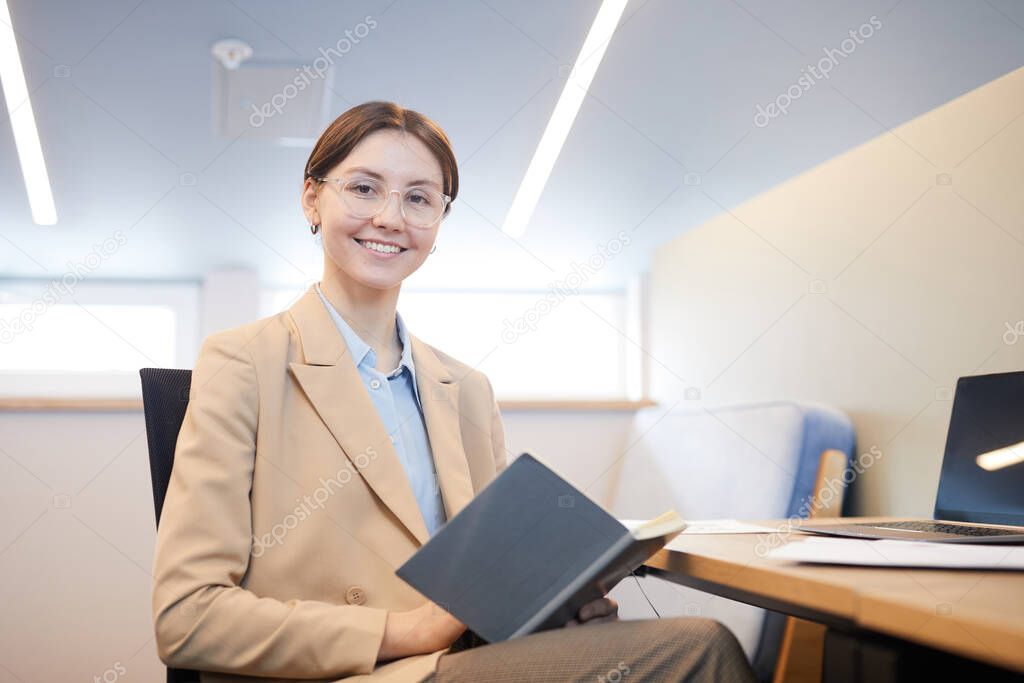 Portrait of elegant young businesswoman wearing glasses and smiling at camera while posing at workplace in minimal office interior, copy space
