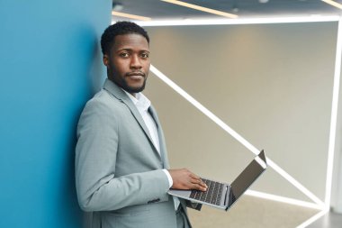 Side view waist up portrait of contemporary African-American businessman holding laptop and looking at camera while standing against blue wall in office, copy space clipart
