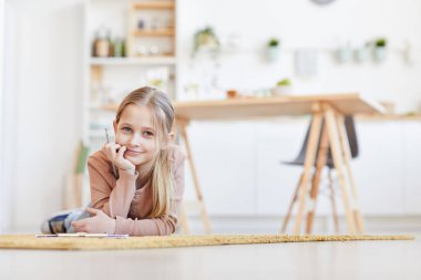 Güzel sarışın kızın sıcak tonlu portresi kameraya gülümseyen ve halı döşemesinde uzanan rahat ev içinde çalışırken ya da çalışırken,