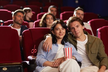 Sinemada film izleyen bir grup insan, kucaklaşan ve kameraya bakan gülümseyen genç çifte odaklanırken ön sırada kırmızı kadife koltuklarda oturuyorlar.