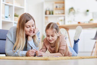 Sıcak tonda gülümseyen anne ve kızın sıcak bir portresi halının üzerinde yatarken rahat bir evde çizim yapıyor ya da ders çalışıyor.