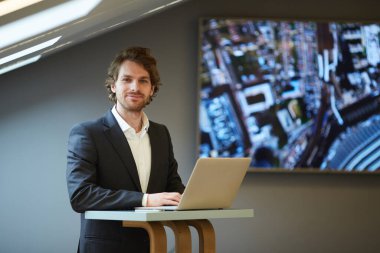 Portrait of handsome bearded businessman looking at camera and smiling while posing at standing table in futuristic office interior with city map in background, copy space clipart