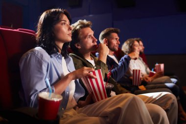 Sinemada film izleyen ve kırmızı koltuklarda otururken patlamış mısır yiyen bir grup arkadaşın yan görüntüsü.