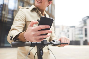 Trençkotlu neşeli bir gencin düşük açılı portresi akıllı telefon kullanırken ve arka planda şehir binalarıyla elektrikli scooter sürerken başka yöne bakıyor.