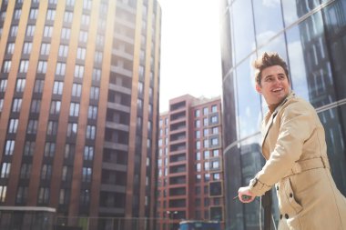 Trençkotlu neşeli bir gencin geniş açılı portresi. Arka planda kentsel şehir binaları ve arka planda hızlı bir şekilde yürürken arkasına bakıyor.