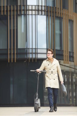 Şehir merkezinde işe gidip gelirken elektrikli scooter 'ın yanında yürüyen neşeli genç iş adamının dikey boy portresi.