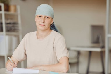 Başörtüsü takmış, doktor muayenehanesinde evrakları doldururken kameraya bakan kel bir kadının sıcacık belli portresi, kanser farkındalığı, kopyalama alanı