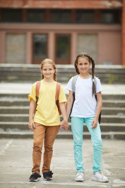 Okul binasına karşı poz verirken sırt çantalı iki sevimli kızın dikey boy portresi.