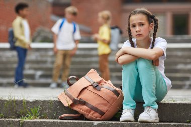 Full length portrait of sad schoolgirl looking at camera while sitting on stairs outdoors with group of children in background, copy space clipart