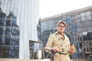 Trençkot giyen, elinde kahve fincanı tutarken kameraya bakan ve arka planda cam binalar olan şehir merkezinde elektrikli mobilete yaslanan modern genç adamın belden yukarı portresi.