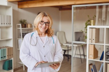 Modern klinikte çalışırken kameraya gülümseyen ve panoyu tutan olgun kadın portresi.