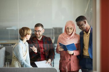 Ofisin içinde cam duvarın arkasında dikilirken iletişim kuran ve el kol hareketi yapan neşeli çoklu etnik iş ekibinin belini kaldırın.