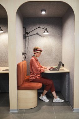 Ofisteki kafe kabininde çalışırken dizüstü bilgisayar kullanan dövmeli genç bir kadının tam boy yan görünüm portresi, boşluğu kopyala