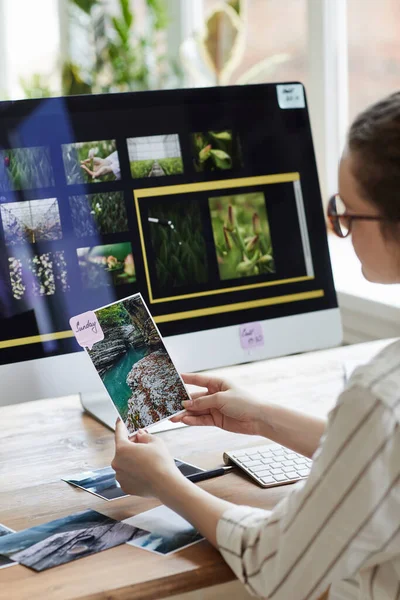 年轻女性摄影师坐在电脑屏幕上的照片编辑软件桌边 一边举着照片一边微笑的垂直肖像 — 图库照片