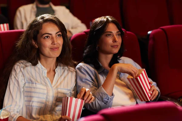 Sinemada film izleyen ve kırmızı kadife koltuklarda oturup patlamış mısır yiyen iki genç kadının portresi.